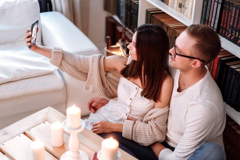 A happy couple taking a selfie indoors, surrounded by candles and books, creating a cozy atmosphere.