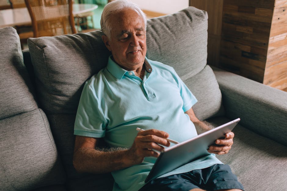 Relaxed elderly man using a tablet with a stylus, seated on a comfy sofa indoors.