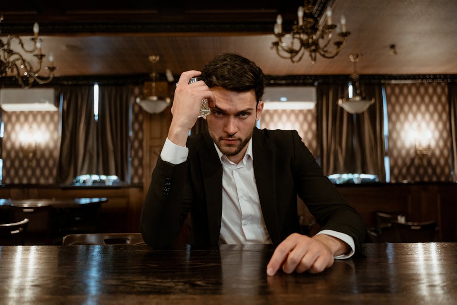 A serious man in a suit sits in a dimly lit restaurant holding a glass, exuding mystery and sophistication.