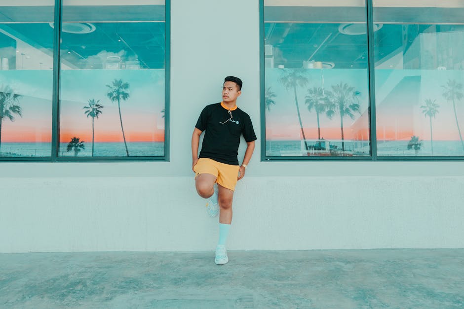 Full body of confident ethnic male in activewear leaning on wall with bog windows decorated with pictures of tropics and looking away