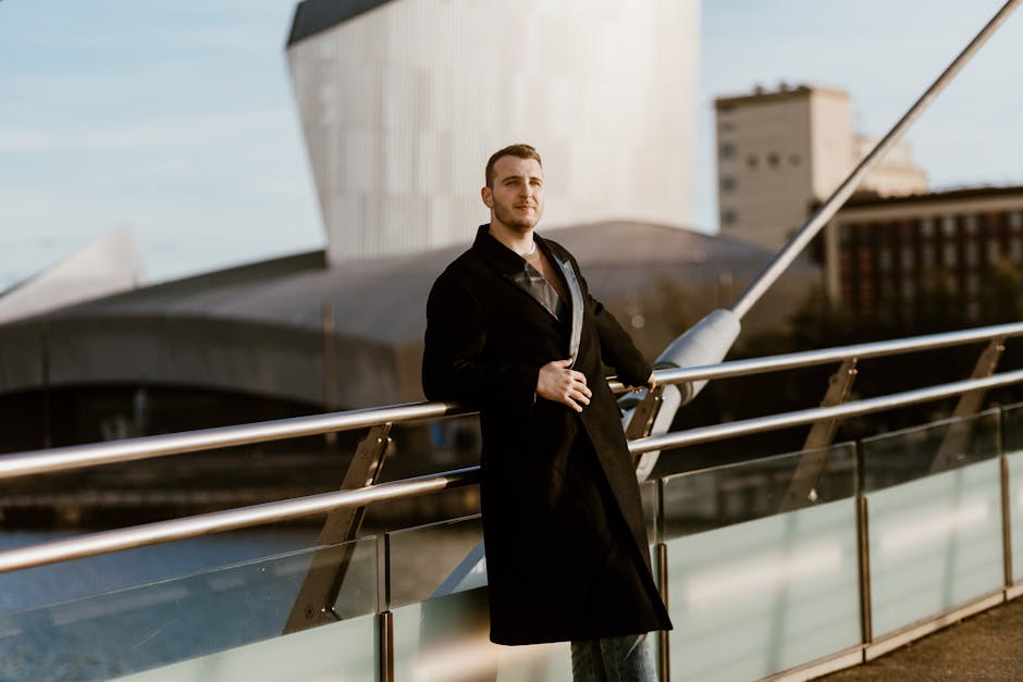 A fashionable man poses on a bridge in an urban setting with modern architecture.