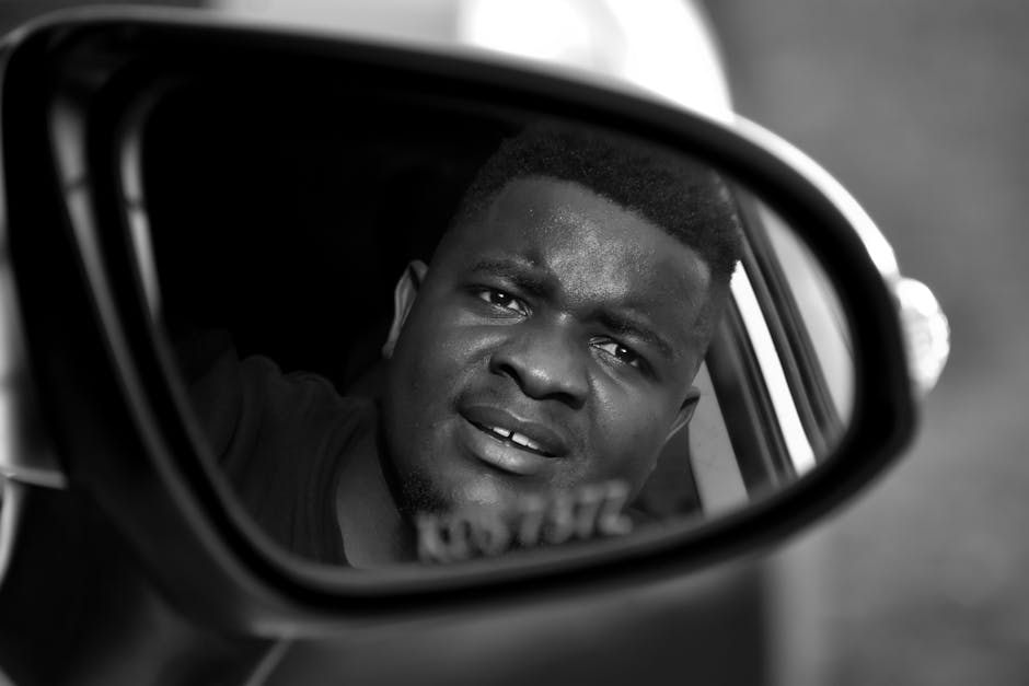 A black and white image of a man captured through a car side mirror, conveying a thoughtful expression.