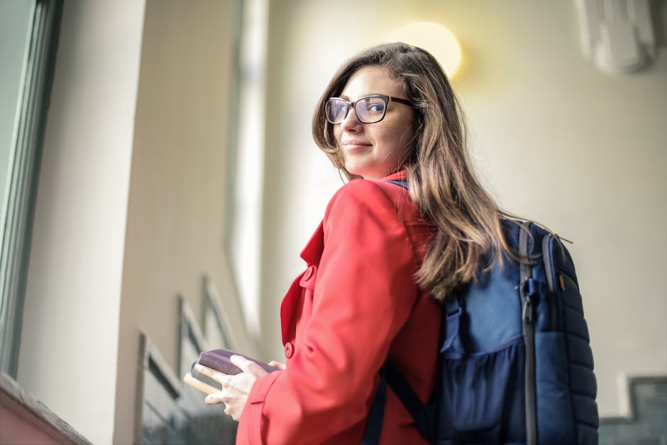 Smiling woman wearing eyeglasses and red coat holds books while standing indoors.