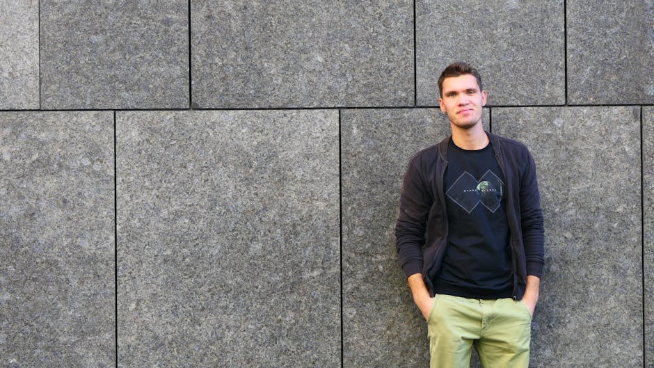 Man in casual attire leans against a stone wall, exuding a relaxed vibe.