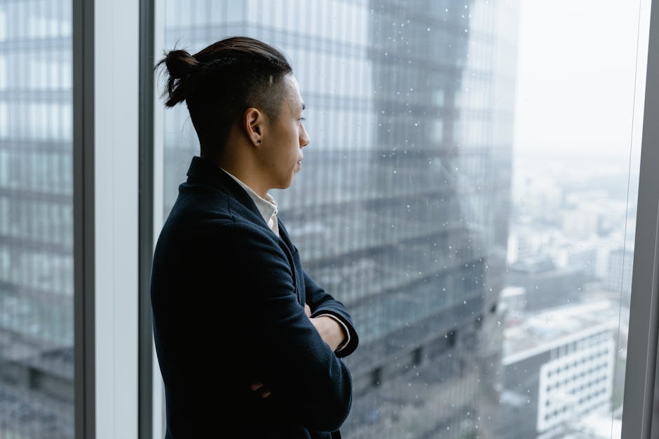 Asian businessman in formal attire looks out from office window on a rainy day.