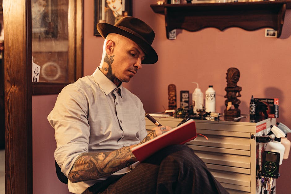 A man with tattoos and a hat writes in a notebook in a cozy, artistic studio setting.