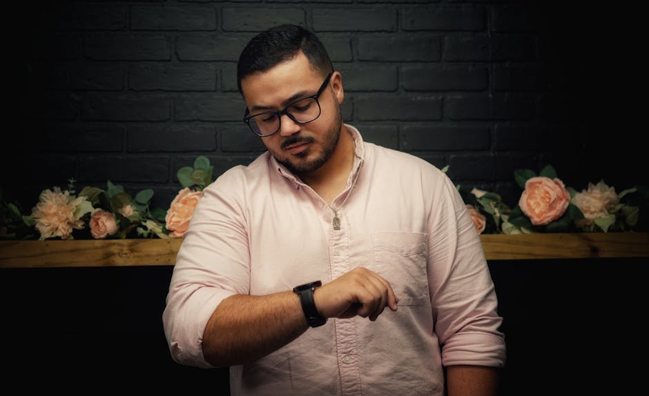 Young man with glasses checking time on wristwatch against black brick wall indoors.